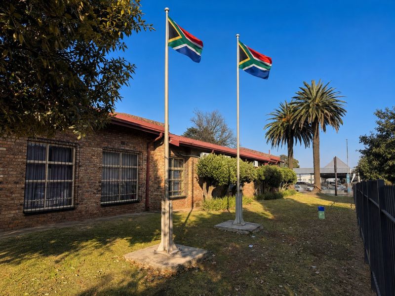 Exterior view of a commercial property featuring a face brick facade, security window bars, and prominent flagpoles set within a landscaped garden area.