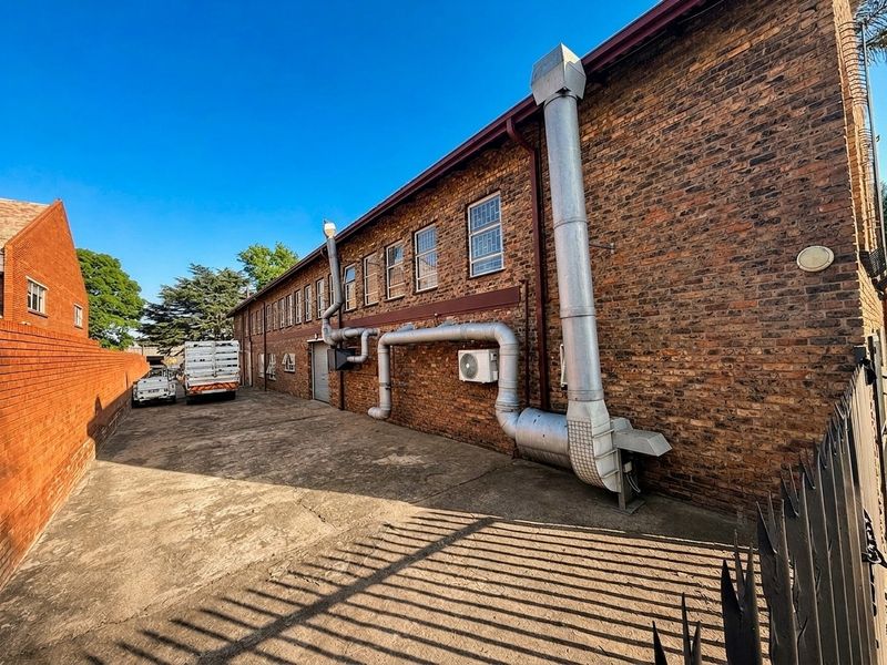 Rear exterior view of a commercial brick building featuring industrial ventilation systems, concrete driveway, and secure perimeter fencing.