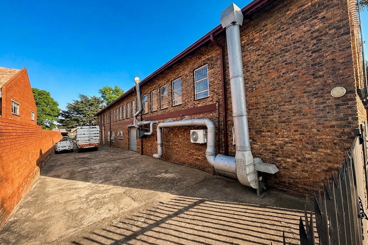 Rear exterior view of a commercial brick building featuring industrial ventilation systems, concrete driveway, and secure perimeter fencing.