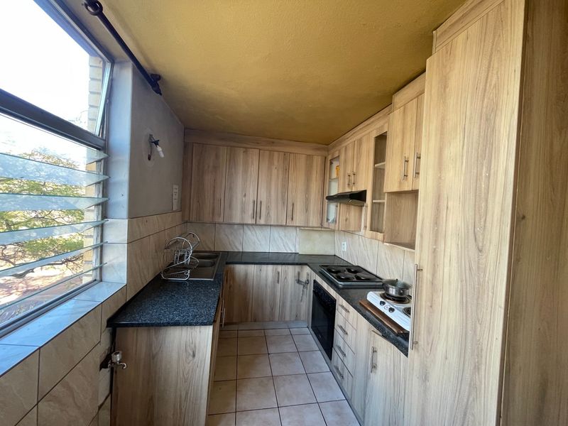 A compact kitchen featuring wood-look laminate cabinetry, granite countertops, built-in oven, and hob, with tiled finishes and natural light from large louvre windows.