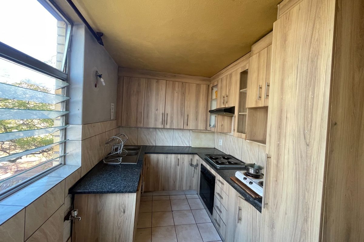 A compact kitchen featuring wood-look laminate cabinetry, granite countertops, built-in oven, and hob, with tiled finishes and natural light from large louvre windows.