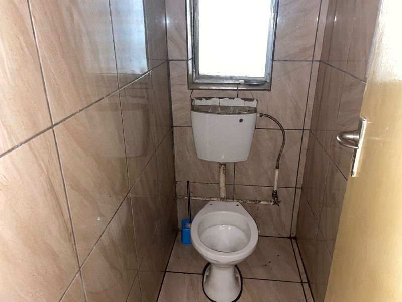 A small, dated bathroom featuring full-height ceramic wall tiling, a standard toilet with an exposed cistern, and a window.