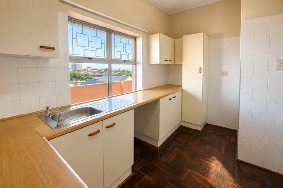 A functional, empty kitchen featuring parquet flooring, laminate countertops, built-in cabinetry, and a tiled splashback with a view through the window.
