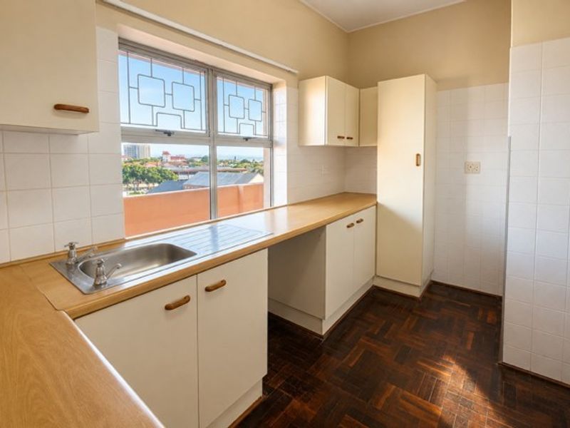 A functional, empty kitchen featuring parquet flooring, laminate countertops, built-in cabinetry, and a tiled splashback with a view through the window.