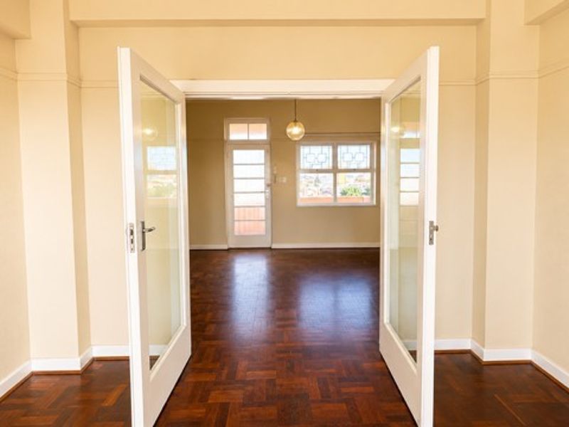 An empty living area featuring classic parquet flooring, neutral walls, and elegant french doors leading to an adjacent room with natural light.