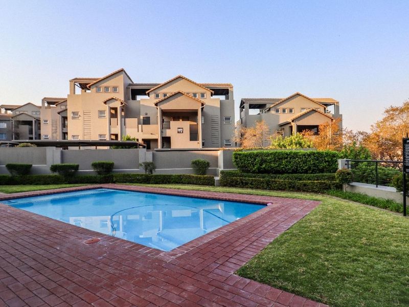 A well-maintained communal swimming pool area featuring brick paving, manicured hedging, and landscaped garden surrounds within a residential apartment complex.