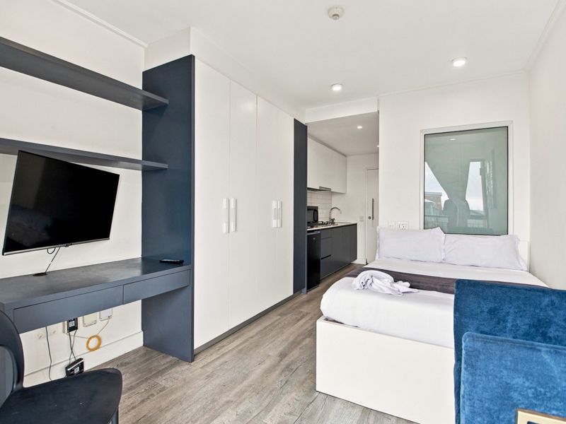 Modern open-plan studio apartment featuring a compact kitchenette, built-in storage cabinetry, a dedicated desk area, and contemporary laminate flooring throughout.