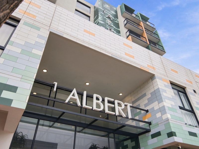 Modern apartment building facade featuring a tiled exterior, glass entrance canopy, and prominent building signage at the main entrance.