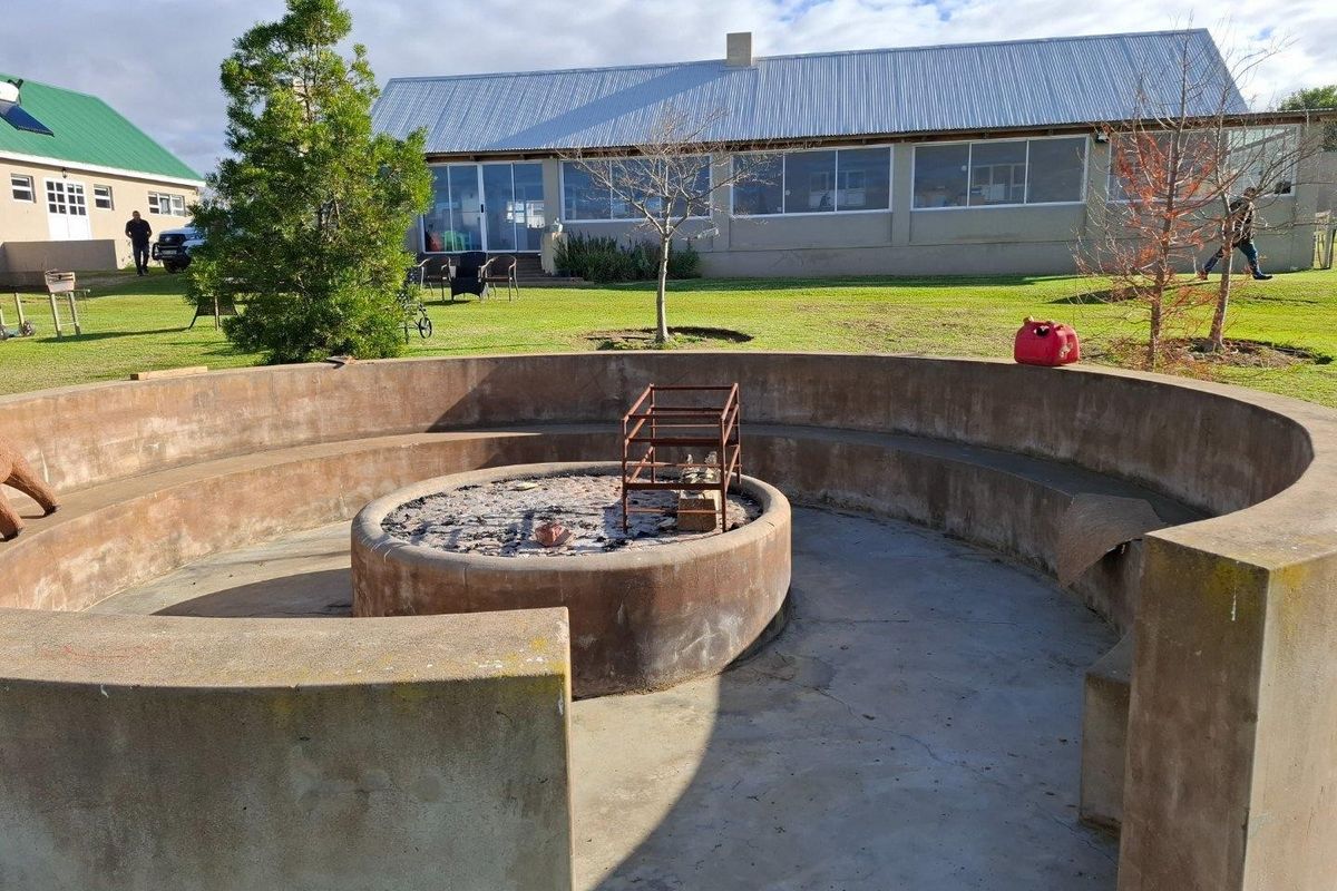 Outdoor garden area featuring a circular built-in concrete fire pit with integrated bench seating, set against the backdrop of a farm-style building.