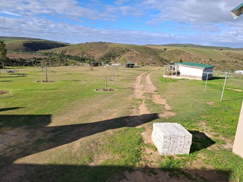 Expansive farm garden area featuring a dirt driveway, established trees, and a small outbuilding set against a scenic hilly landscape.