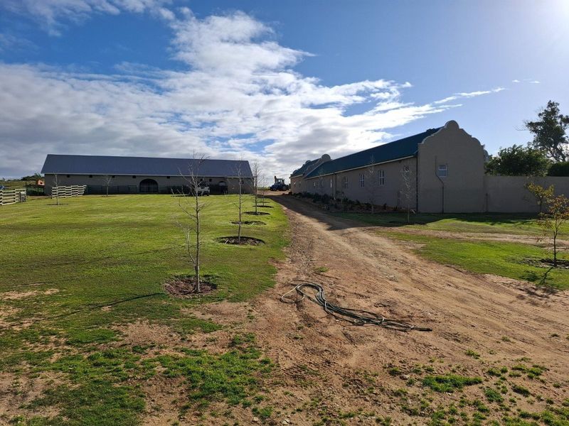 A view of the farm property featuring large outbuildings with metal roofing, a dirt driveway, and expansive lawn areas.