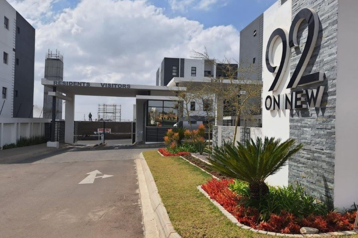 Modern apartment complex entrance featuring stone-clad signage, paved driveway, and landscaped garden beds at the secure access point.