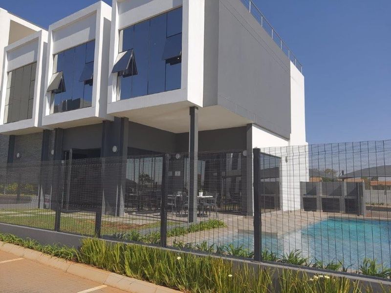 Modern apartment building exterior featuring a swimming pool, paved patio area, and secure mesh perimeter fencing.