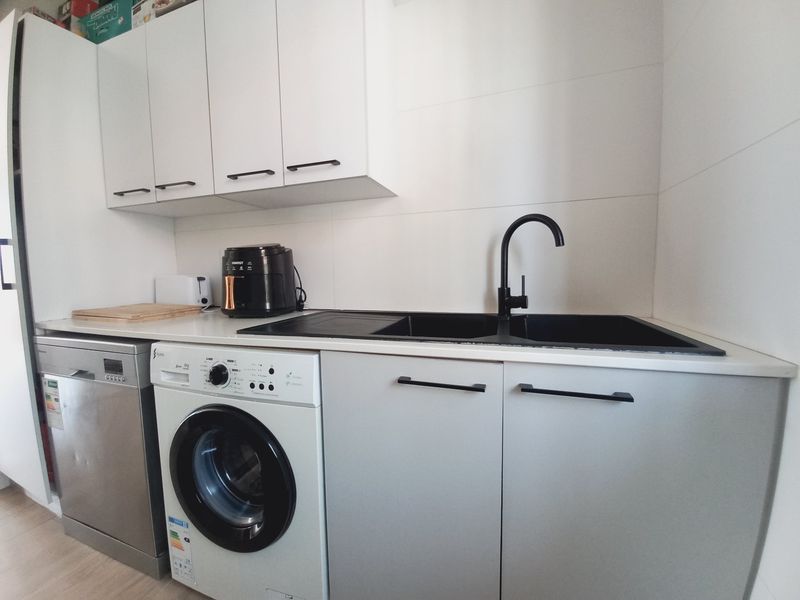 A compact, modern kitchen featuring white upper cabinetry, grey lower cabinets, a black sink with matching tap, and integrated laundry and dishwasher appliances.