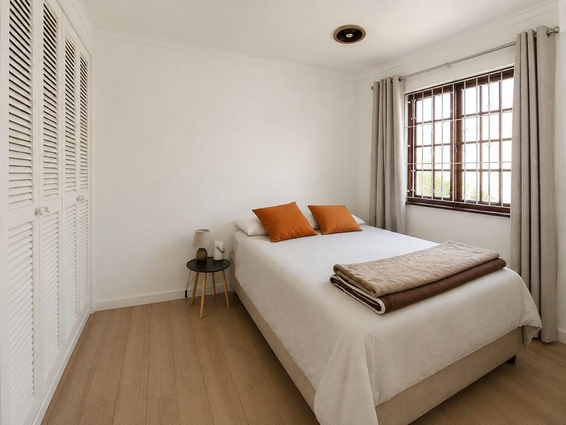A well-maintained bedroom featuring light laminate flooring, built-in louvered cupboards, and a window fitted with security bars.