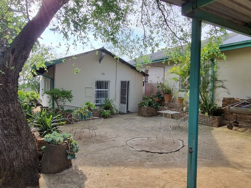 A covered outdoor patio area featuring concrete flooring, brick planter boxes, and security bars on the windows, set within a garden environment.