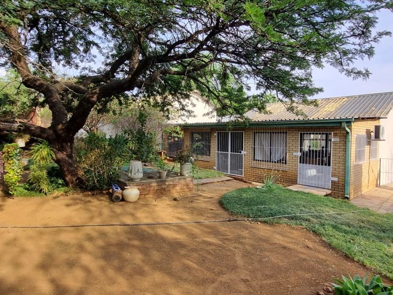 Rear view of a face brick small holding dwelling featuring a corrugated metal roof and security fixtures, set within a mature garden landscape.