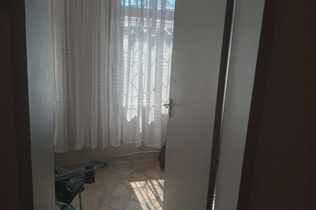 A simple bedroom space featuring tiled flooring and a window with security bars, viewed through an open doorway.