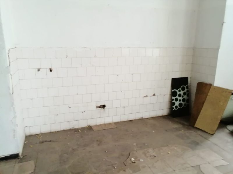 An empty, dilapidated kitchen area featuring damaged wall and floor tiles with exposed plumbing connections, requiring significant renovation and repair.