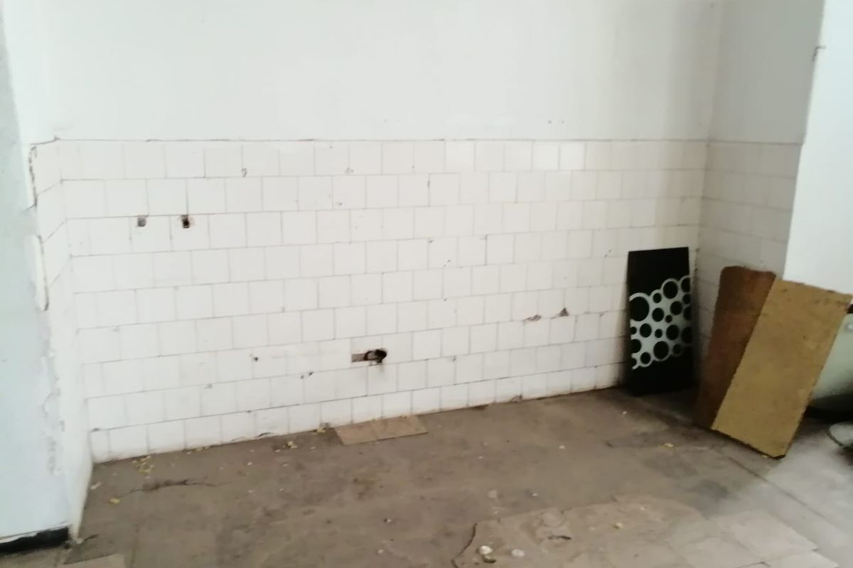 An empty, dilapidated kitchen area featuring damaged wall and floor tiles with exposed plumbing connections, requiring significant renovation and repair.
