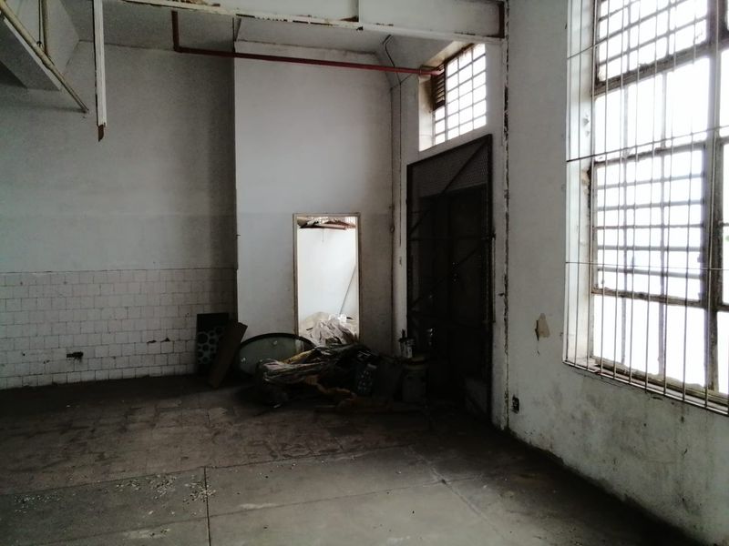 An industrial utility space featuring high ceilings, exposed fire piping, large steel-framed windows with security grilles, and concrete flooring in need of refurbishment.