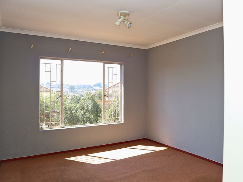 A vacant bedroom featuring grey walls, carpeted flooring, and a large window with security bars, offering a bright and neutral space.