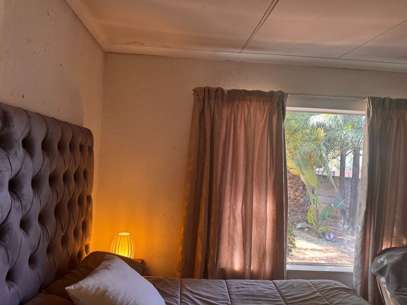 A bedroom featuring a window with garden views, showing signs of ceiling water damage and wall wear requiring maintenance.
