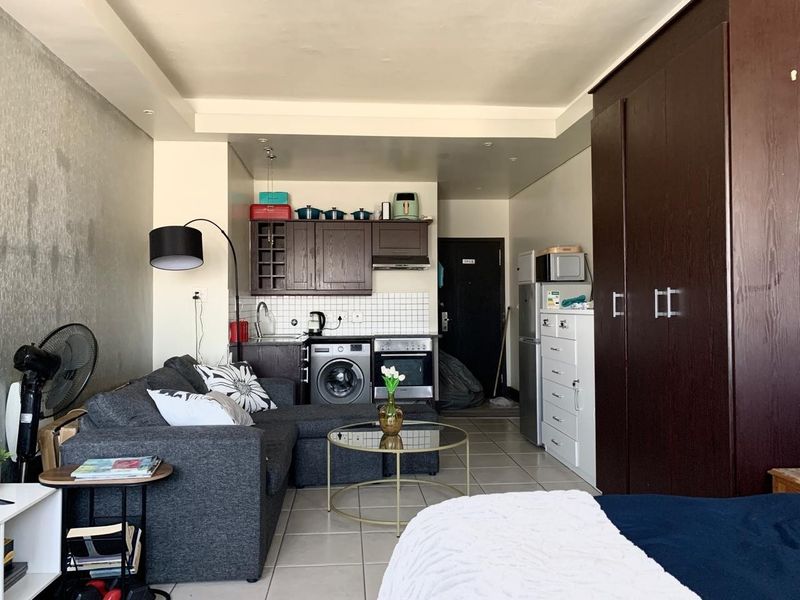 Compact open-plan studio apartment featuring a kitchenette with built-in cabinetry, oven, and hob, alongside a tiled living and sleeping area with built-in storage.