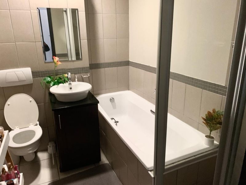 A functional bathroom featuring a built-in bathtub, wall-mounted toilet, and a vanity cabinet with a ceramic basin, finished with neutral wall and floor tiles.