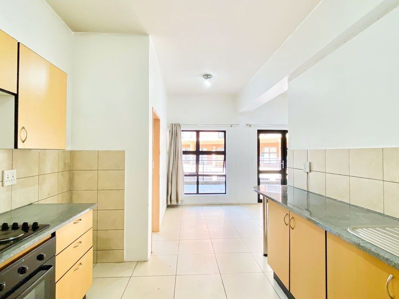 A functional, empty kitchen featuring light wood-finish cabinetry, tiled backsplash, built-in oven and hob, and tiled flooring, leading into an open-plan living area.