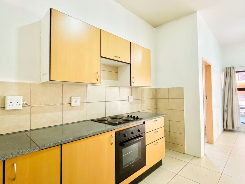 A functional kitchen featuring light wood-finish cabinetry, dark countertops, a built-in electric hob and oven, and neutral tiled backsplash and flooring.