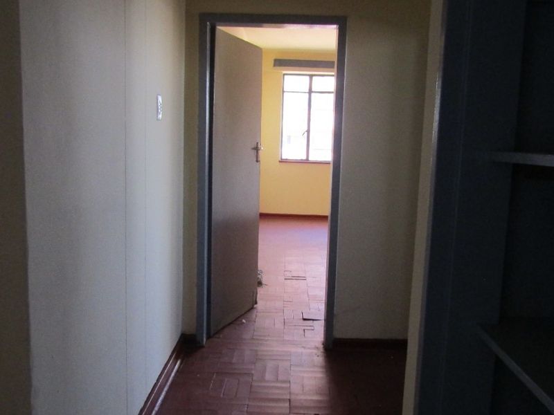 A narrow hallway featuring worn parquet flooring leading into an adjacent room with natural light.