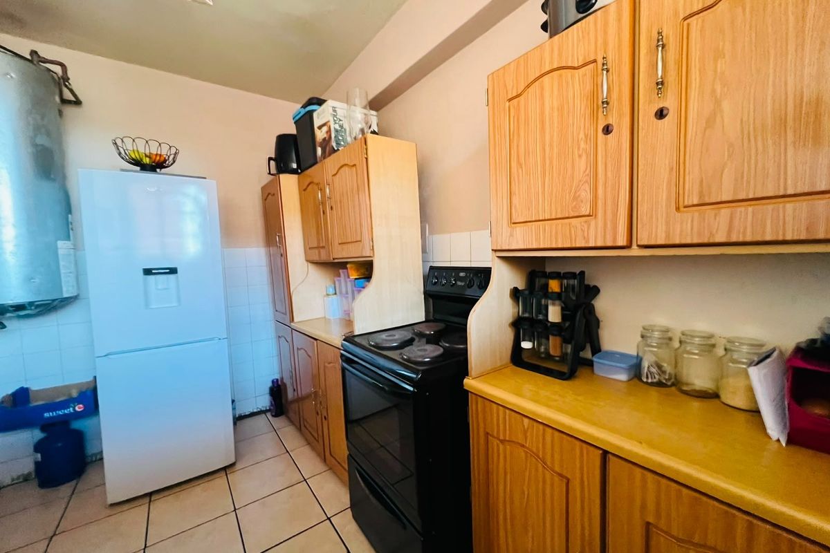 A functional kitchen featuring wooden cabinetry, tiled flooring, a tiled backsplash, and a built-in electric stove with an exposed geyser.