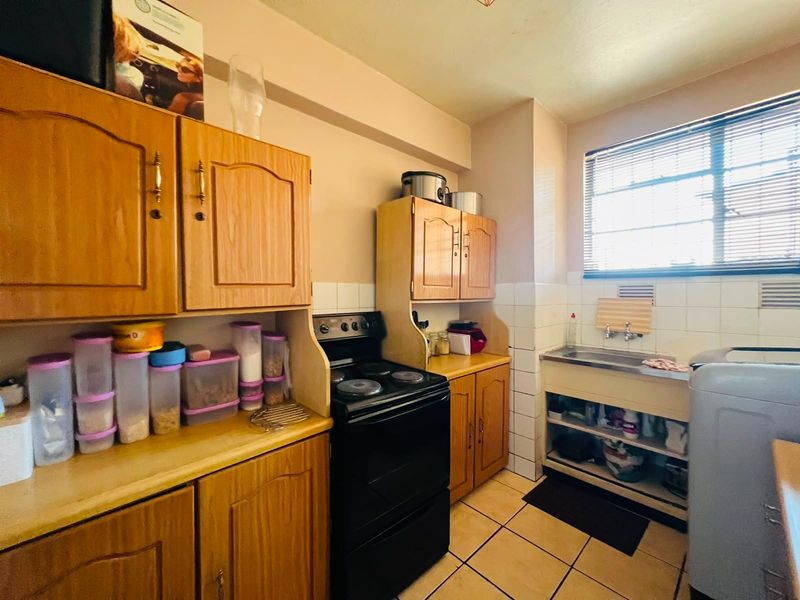 A compact kitchen featuring wooden cabinetry, a built-in electric stove, tiled backsplash, and a stainless steel sink with tiled flooring.