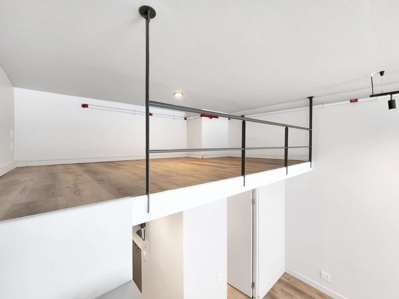 A bright and modern mezzanine loft area featuring wood-look laminate flooring, a sleek metal safety railing, and exposed industrial-style fire sprinkler piping.