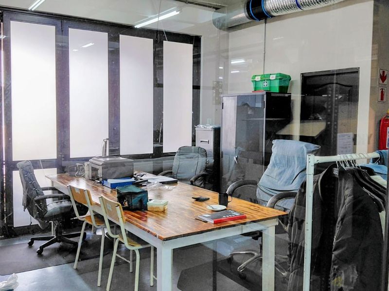 A functional industrial office space featuring glass partitioning, exposed ventilation ducting, and overhead fluorescent lighting, suitable for administrative or operational use.