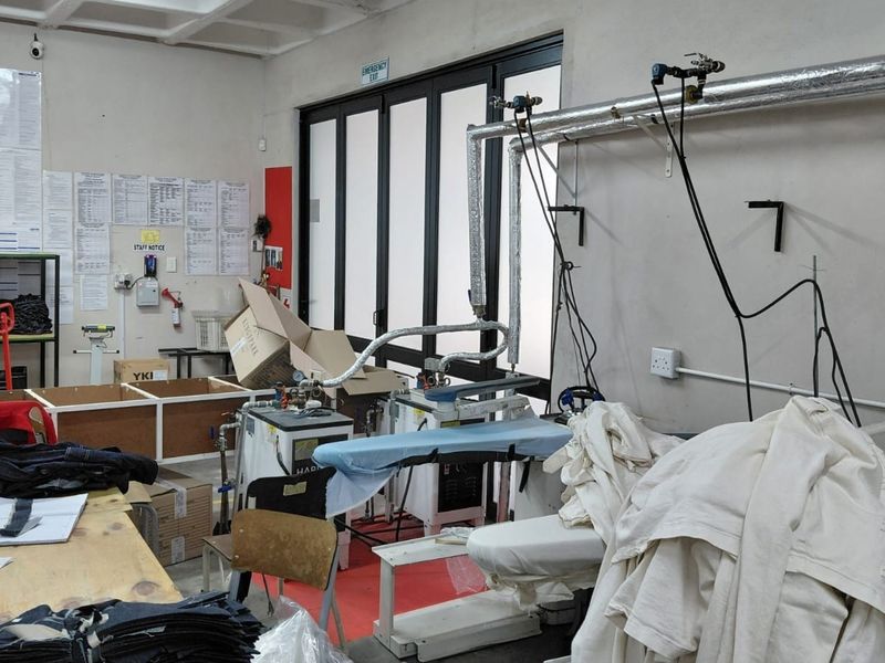 Industrial utility space featuring fixed steam pressing equipment, exposed overhead piping, and an emergency exit door within a functional work environment.