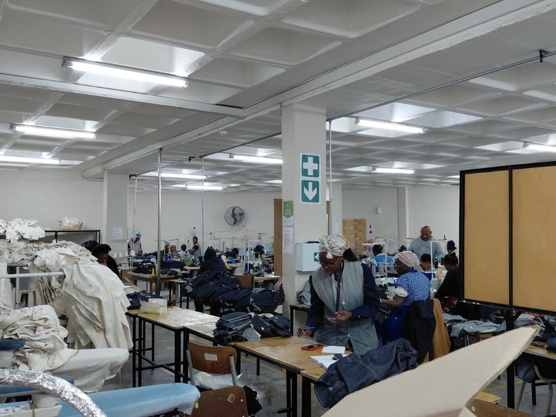 A large industrial workspace featuring a coffered ceiling, overhead fluorescent lighting, and structural support columns, currently utilized as a garment manufacturing facility.