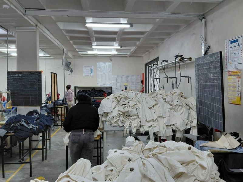 Industrial manufacturing workspace featuring exposed concrete ceilings, fluorescent lighting, and functional utility infrastructure for garment production.