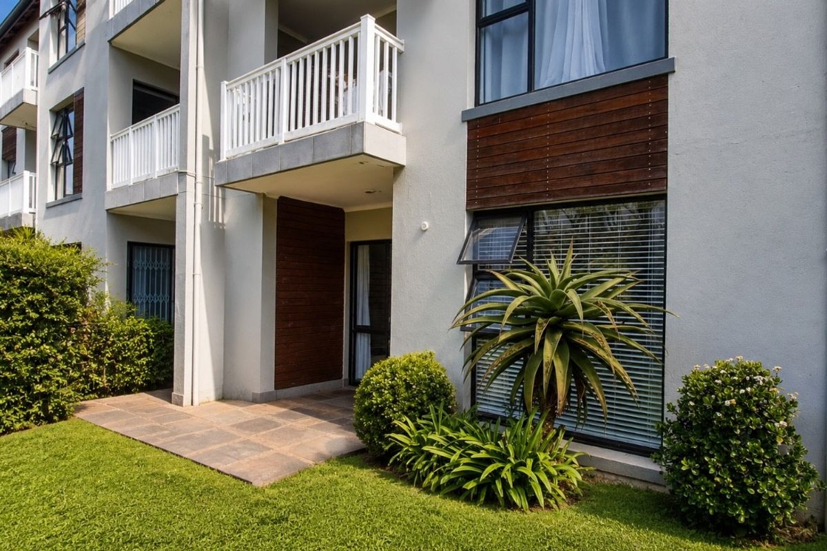 Ground floor apartment exterior featuring a paved patio area, timber cladding accents, and a private balcony above, set within a well-maintained garden.