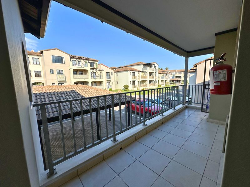 A covered balcony featuring tiled flooring and a metal balustrade, overlooking the apartment complex parking area.