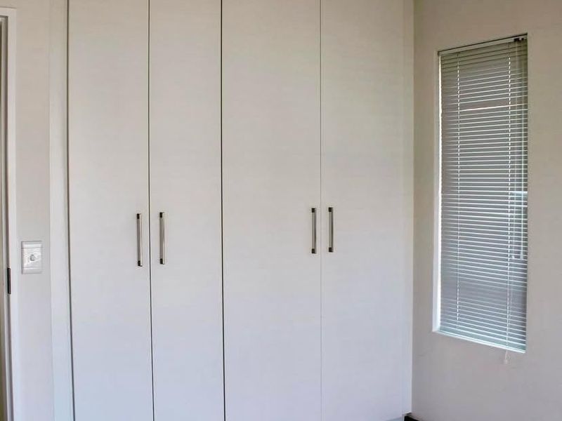 A clean, neutral bedroom featuring floor-to-ceiling built-in cupboards, tiled flooring, and a window fitted with venetian blinds.