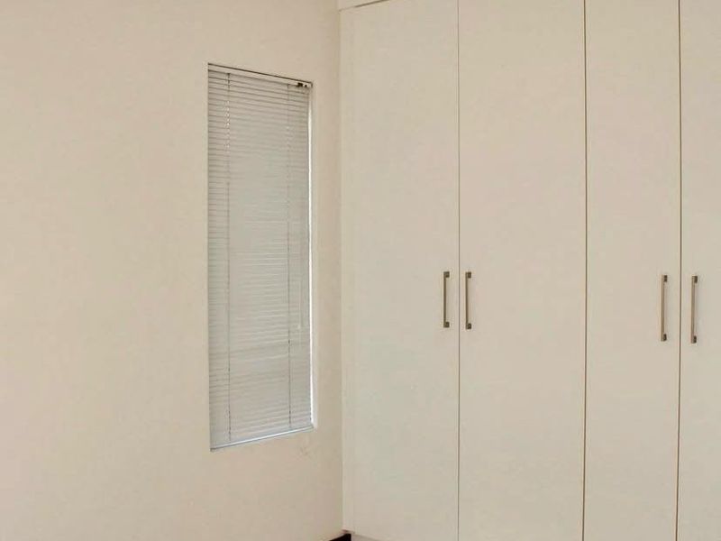 A clean, empty bedroom featuring neutral tiled flooring, built-in floor-to-ceiling cupboards, and a window fitted with venetian blinds.