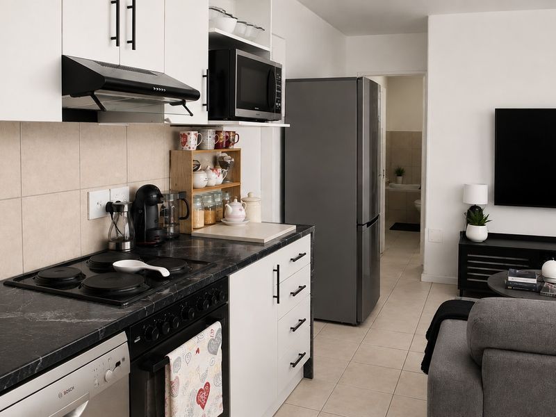Modern open-plan kitchen and living area featuring sleek white cabinetry, dark stone-look countertops, and integrated appliances, flowing into a comfortable lounge space.