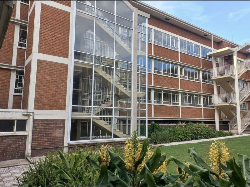 A multi-story commercial building featuring a prominent glass-enclosed staircase, brick facade, and external steel fire escape stairs set against a landscaped garden area.