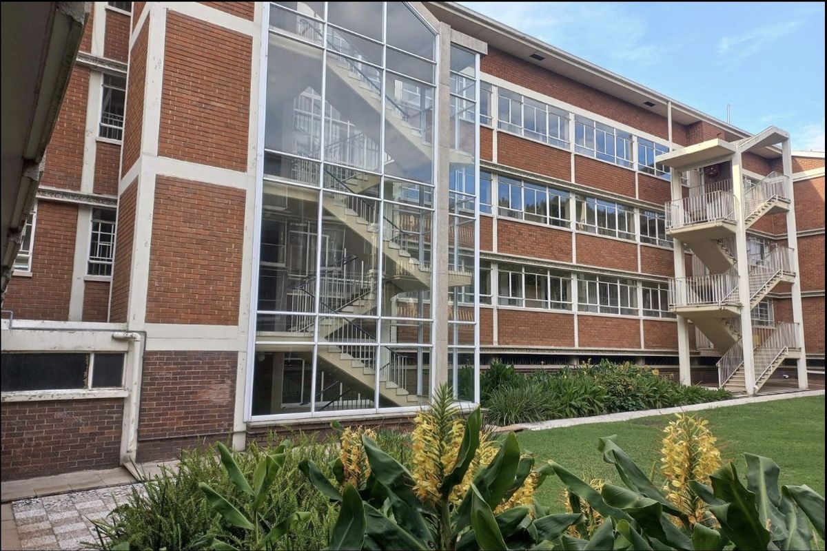 A multi-story commercial building featuring a prominent glass-enclosed staircase, brick facade, and external steel fire escape stairs set against a landscaped garden area.