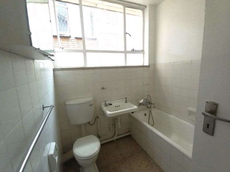 A dated bathroom featuring a standard bathtub, wall-mounted basin, and toilet with white wall tiling and a large window.