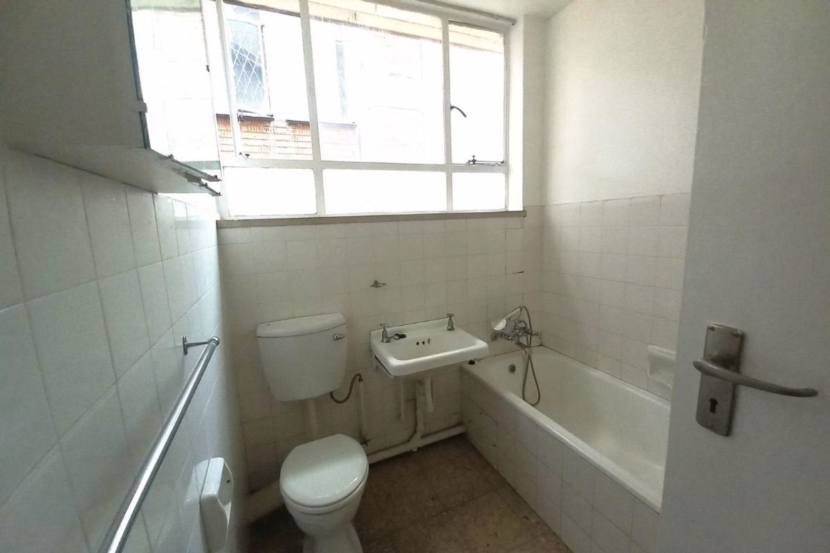A dated bathroom featuring a standard bathtub, wall-mounted basin, and toilet with white wall tiling and a large window.