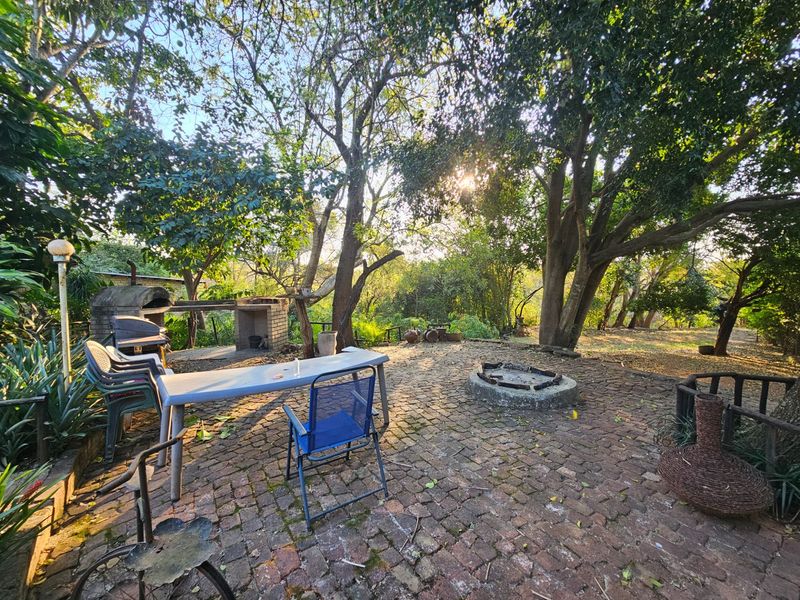 A rustic outdoor garden area featuring a brick-paved patio, a built-in brick braai, and a stone fire pit surrounded by mature trees.