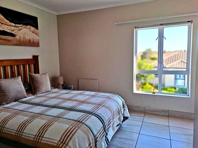 A well-lit bedroom featuring neutral wall tones, tiled flooring, and a large window providing natural light and a view of the surrounding retirement village.